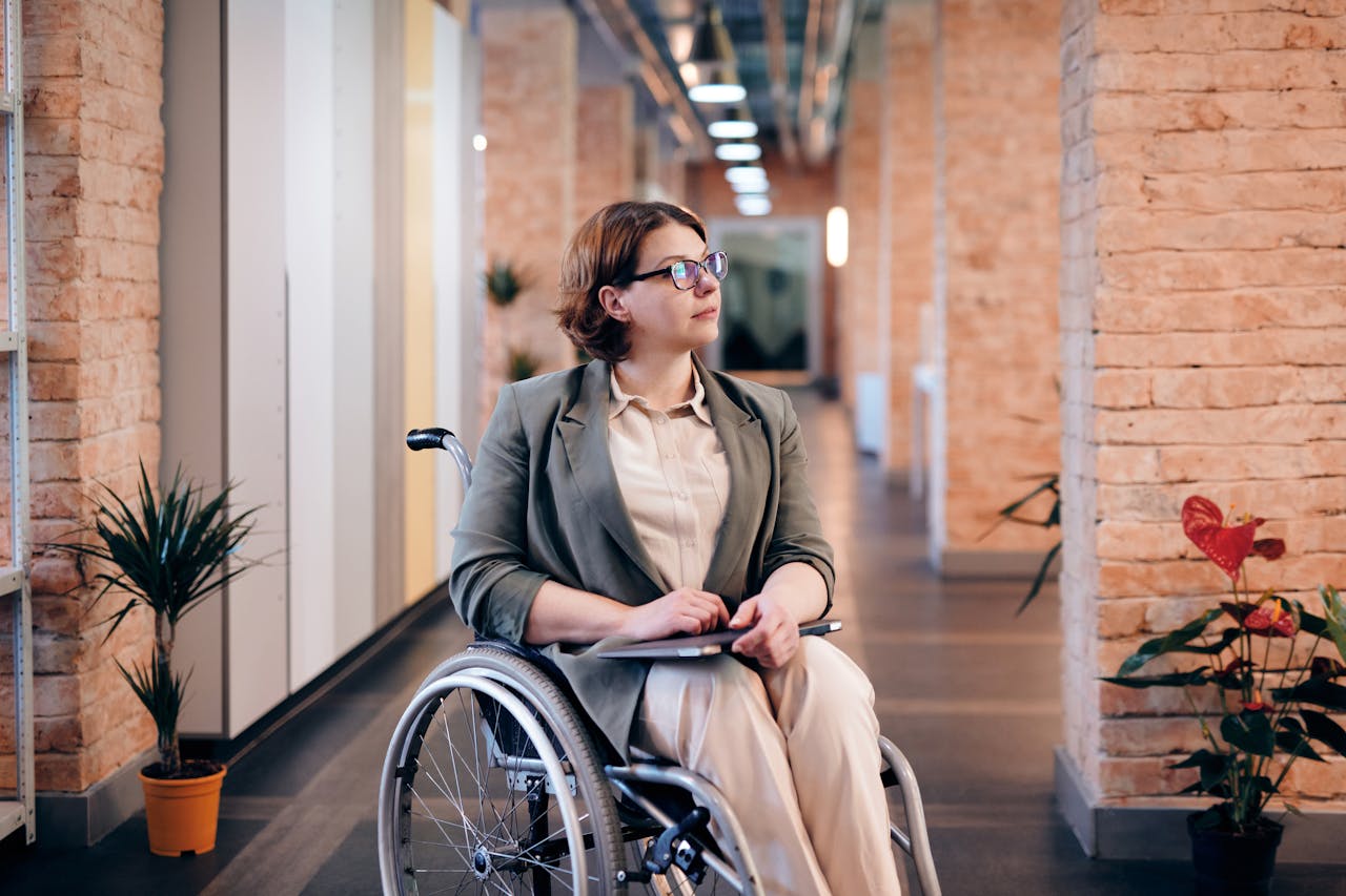 Confident businesswoman in a wheelchair thinking thoughtfully in a modern office hallway.