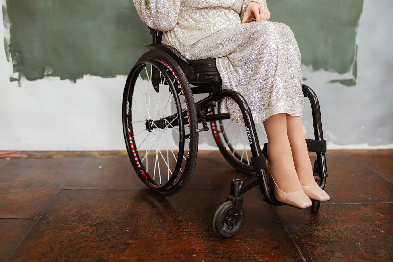 Woman in a silver sequin dress sitting in a wheelchair wearing beige heels, indoors.