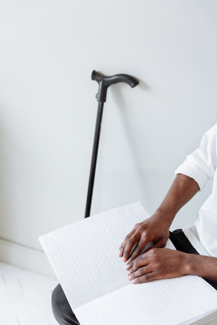An adult reading a Braille book indoors with a cane in the background.